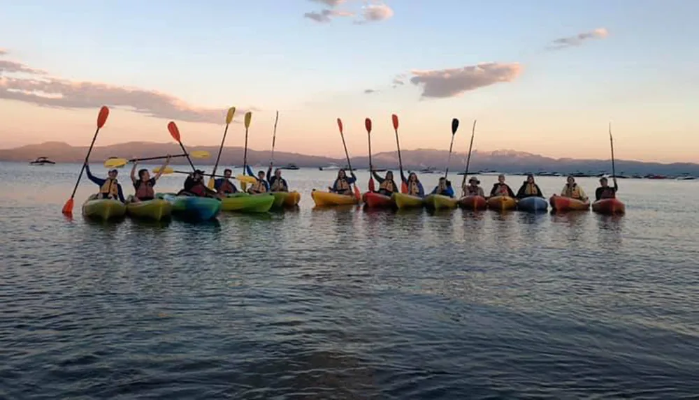 A group of people paddle colorful kayaks in calm waters against a backdrop of a setting sun