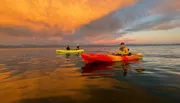 People in colorful kayaks enjoy a serene paddle on calm water under a dramatic orange sunset sky.