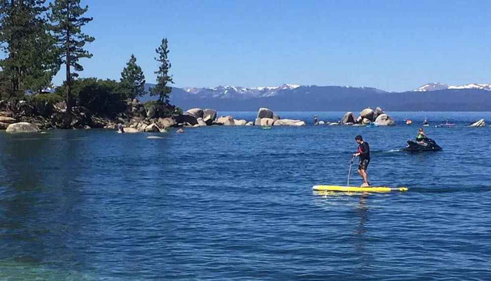 A person is paddleboarding on a clear blue lake with a backdrop of mountains and a jet ski nearby