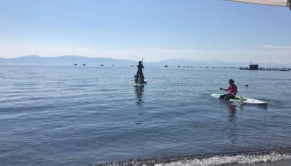 Two people are engaging in water sports one standing on a paddleboard and the other sitting on a kayak on a calm sea with a mountainous horizon in the background
