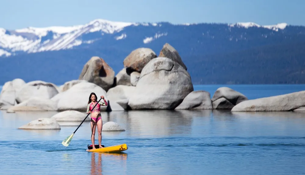 A person is paddleboarding on a calm lake with rocky formations and snow-capped mountains in the background