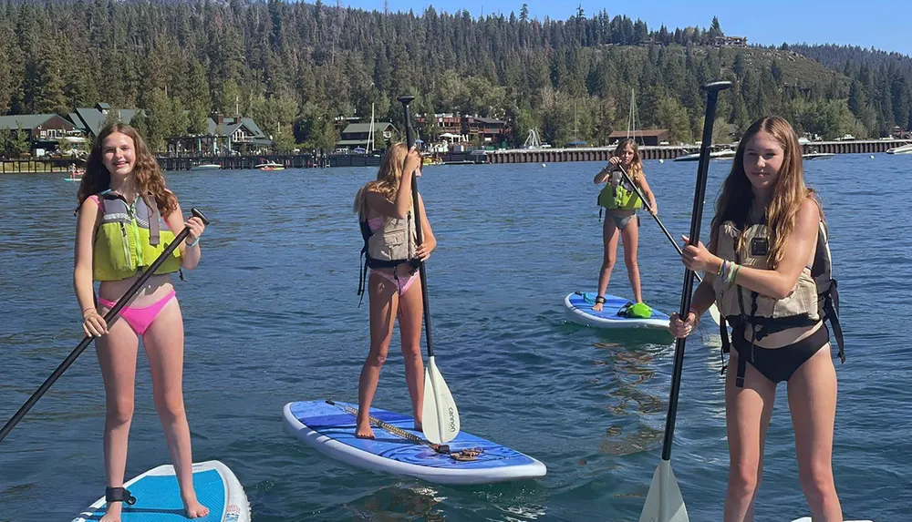 Four people are paddleboarding on a calm lake with a backdrop of a lakeside community and forested hills