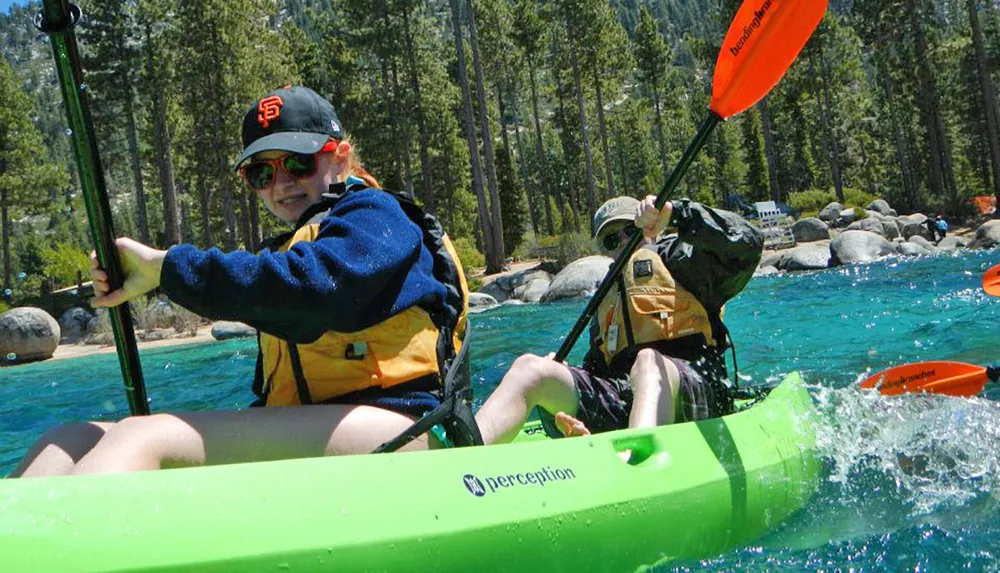 Two individuals are kayaking in a vibrant green kayak on a clear sunny day with a backdrop of trees and rocks