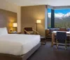 The image shows a neatly arranged hotel room with modern furnishings and a large window offering a stunning view of snow-capped mountains and a pine forest