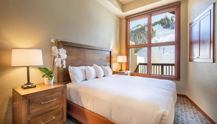 The image shows a cozy bedroom with a large bed wooden furniture and a window offering a view of snowy mountains