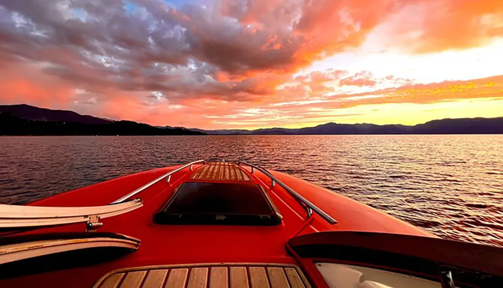 A vibrant red boat is cruising on calm waters with a breathtaking sunset painting the sky in shades of orange and pink behind a silhouette of mountains