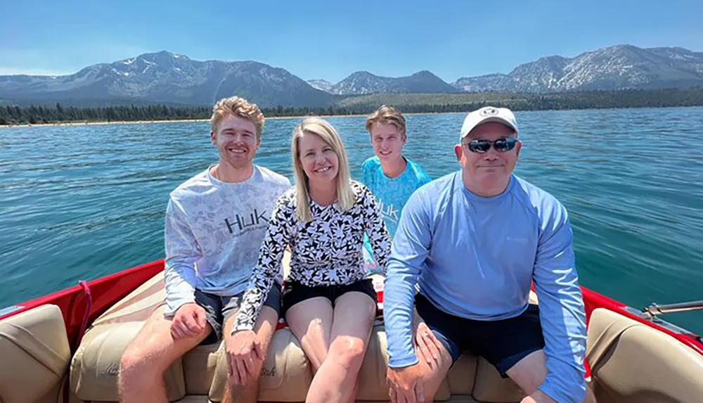 Four people are smiling and posing for a photo while sitting in a boat on a sunny day with a backdrop of clear blue skies and mountains