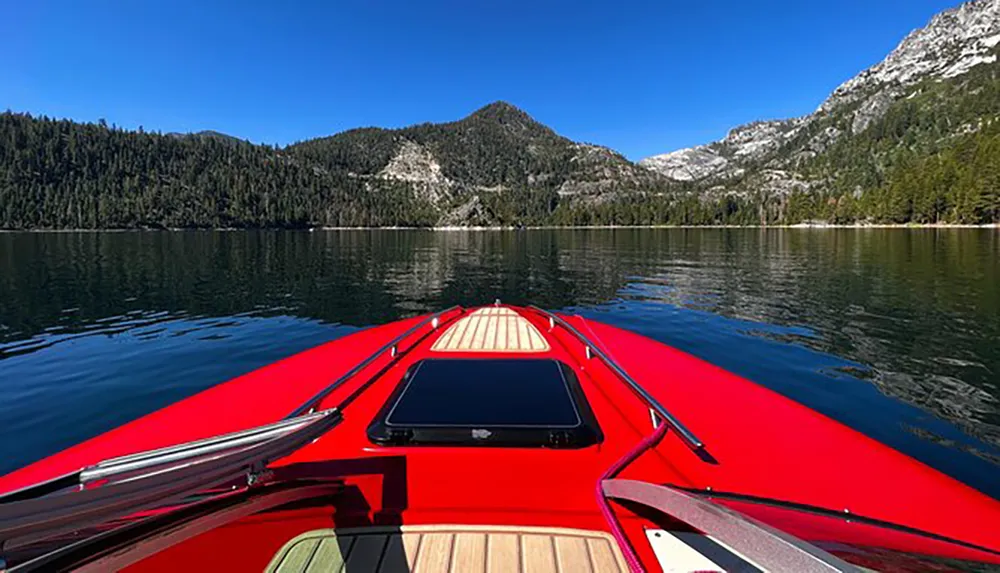 A red boat is cruising on a calm lake with a backdrop of forested mountains under a clear blue sky