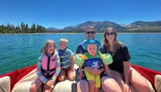 A happy family of five is enjoying a sunny day on a boat with a clear blue lake and mountainous backdrop.