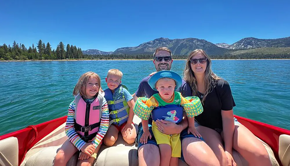 A happy family of five is enjoying a sunny day on a boat with a clear blue lake and mountainous backdrop