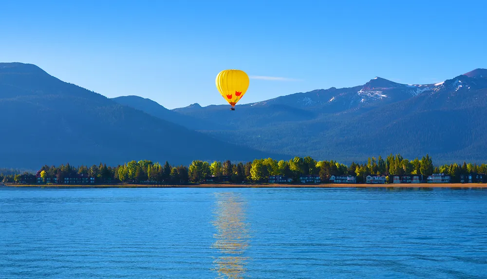 A vibrant yellow hot air balloon floats above a calm blue lake with a mountainous backdrop and a tree-lined shore under a clear sky