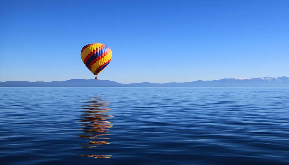 A colorful hot air balloon hovers above a serene blue lake reflecting gently on the waters surface against a backdrop of distant mountains under a clear sky