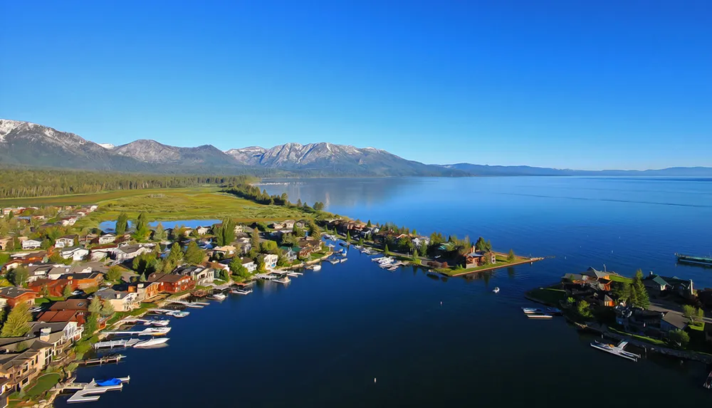 An aerial view of a serene lakeside community with houses dotted along the waterfront boats moored at docks and a backdrop of mountains under a clear blue sky