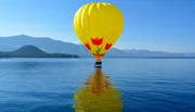 A bright yellow hot air balloon is floating above a serene lake with its reflection on the water and a backdrop of mountains under a clear blue sky.