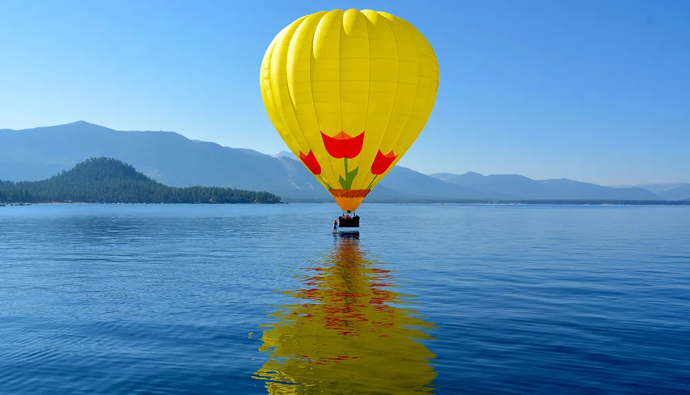 A bright yellow hot air balloon is floating above a serene lake with its reflection on the water and a backdrop of mountains under a clear blue sky