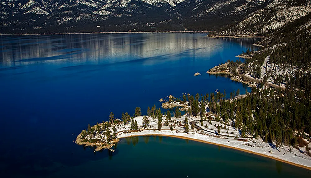 The image shows a serene winter landscape with a deep blue lake surrounded by snow-dusted forests and mountains under a clear sky