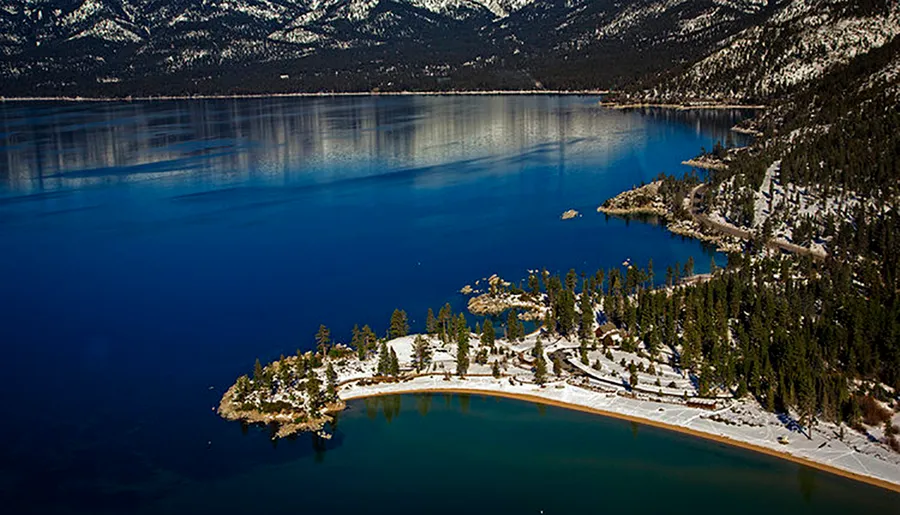 The image shows a serene winter landscape with a deep blue lake surrounded by snow-dusted forests and mountains under a clear sky.