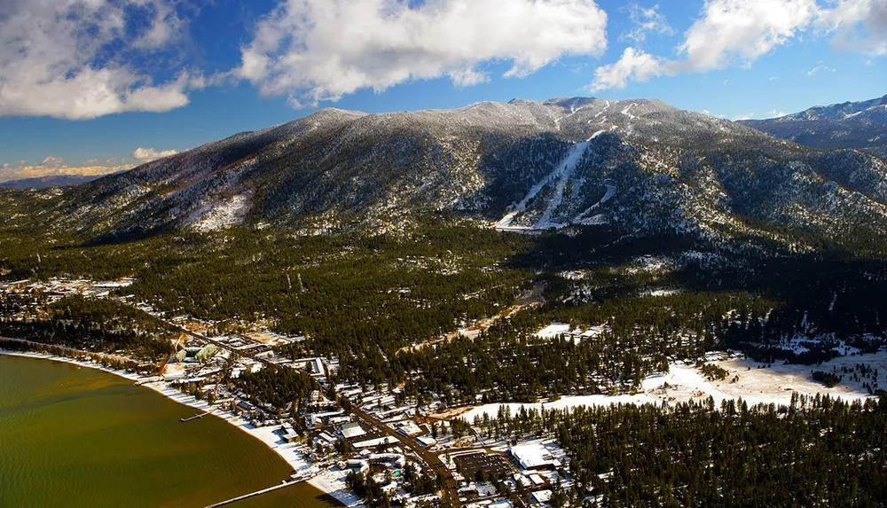 This image shows a snow-covered mountainous landscape with a town nestled at the base near a lake under a partly cloudy sky