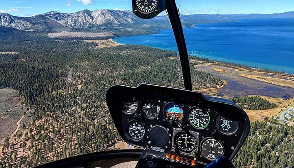 The image shows the view from a helicopter cockpit overlooking a stunning landscape with a lake and mountains in the distance