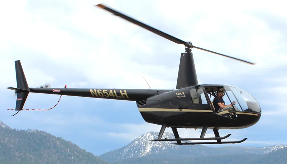A helicopter with its rotor blades in motion is flying over a mountainous area with a pilot visible in the cockpit