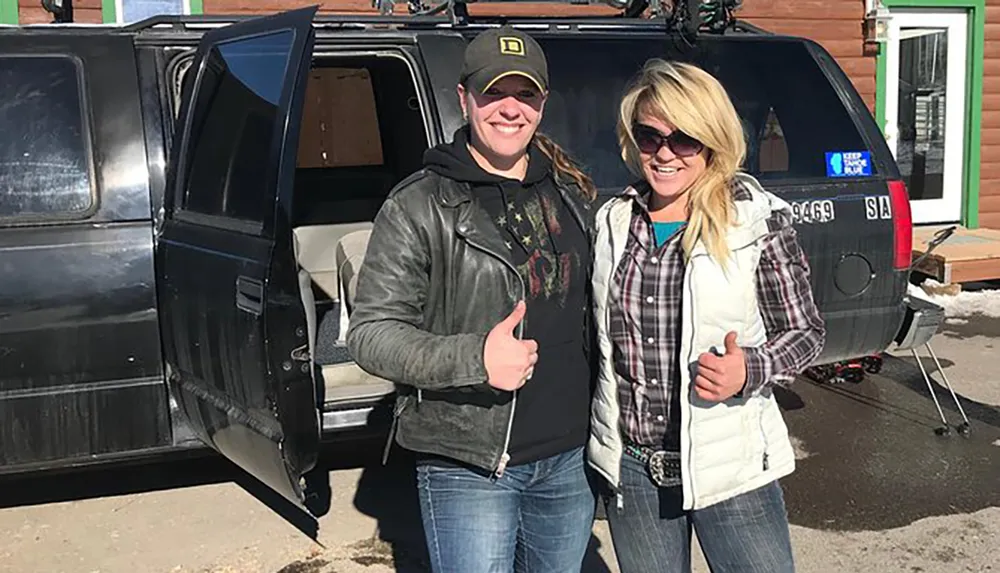 Two people are posing with thumbs up in front of an open-door SUV in a sunny location