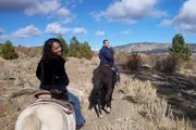 Two individuals are seen horseback riding in a scenic landscape with sparse vegetation under a partly cloudy sky.