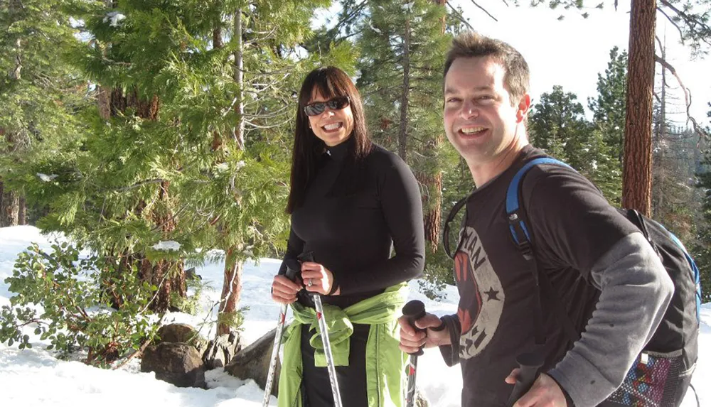 A man and a woman are smiling at the camera while enjoying a sunny day outdoors with snowshoes in a snowy forest setting