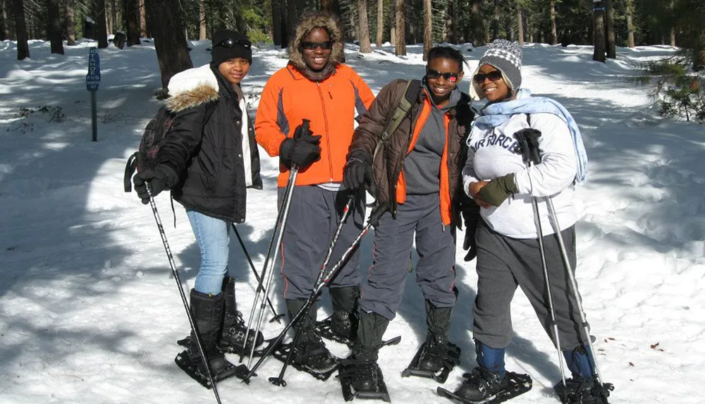 Four individuals are posing for a photo in the snow with snowshoes and poles dressed in winter gear and seem to be enjoying a sunny day of winter sports