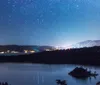 A starry night sky stretches above a serene lake with a small island and distant lights from a lakeside community