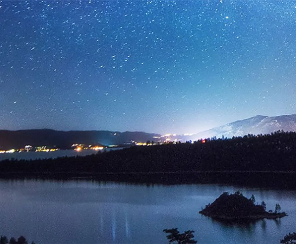 A starry night sky stretches above a serene lake with a small island and distant lights from a lakeside community