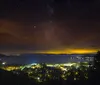 A starry night sky stretches above a serene lake with a small island and distant lights from a lakeside community