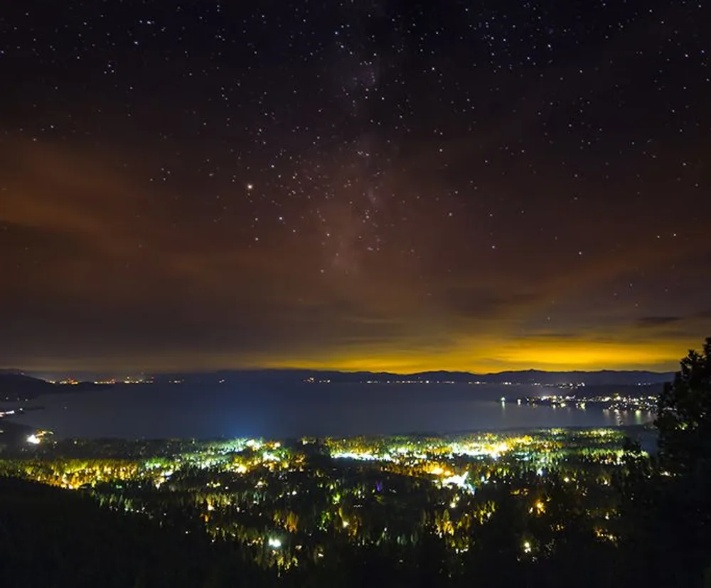 This image features a starry night sky over a brightly lit town adjacent to a large body of water with distant lights reflecting on the waters surface