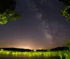 A starry night sky stretches above a serene lake with a small island and distant lights from a lakeside community