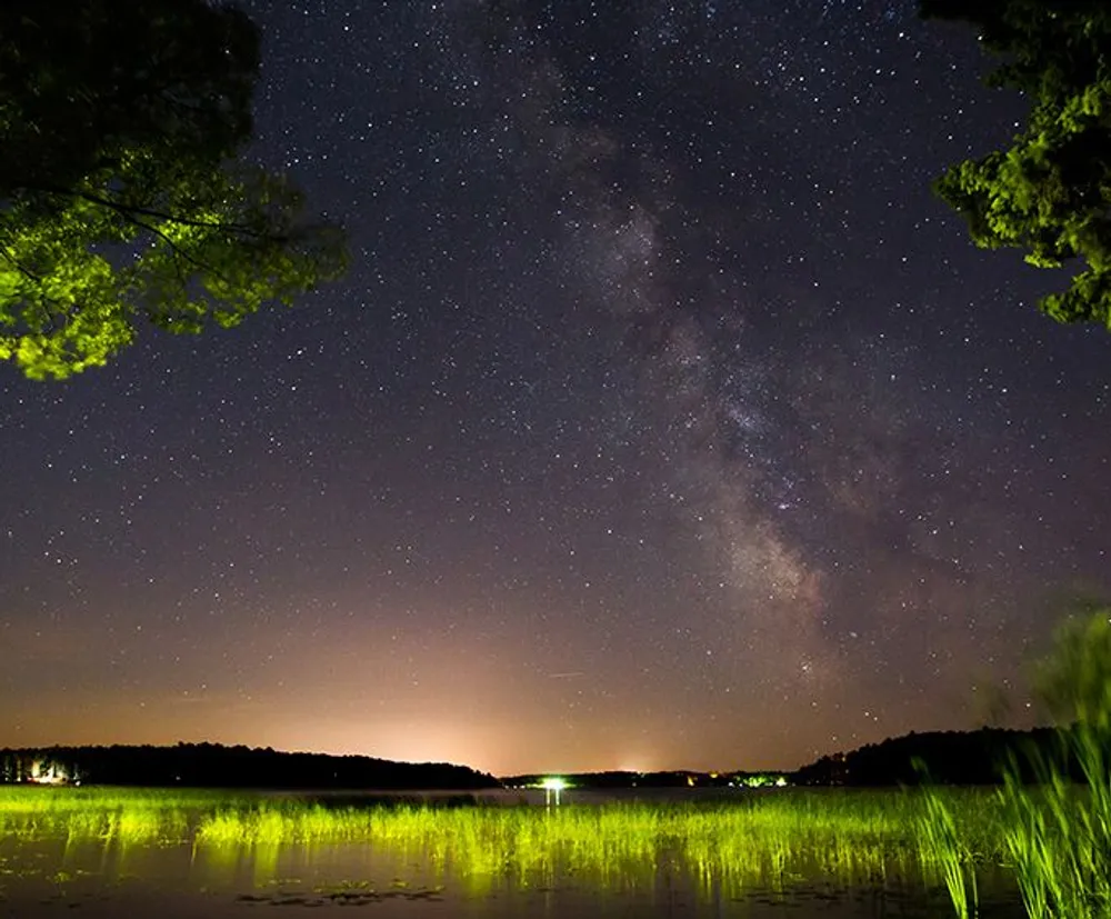 The image captures a stunning night sky filled with stars and the Milky Way with a serene lake in the foreground reflecting a hint of light pollution from a distant source