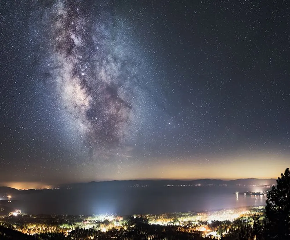 The image showcases a stunning night sky filled with the Milky Way galaxy above a brightly lit cityscape and a large body of water