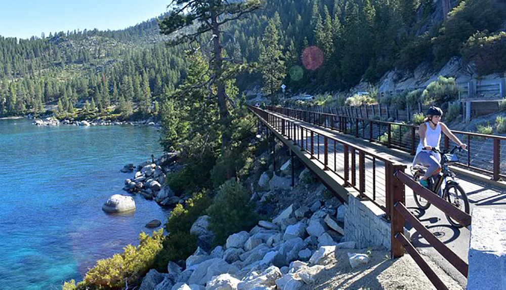 A person is riding a bicycle along a scenic lakeside trail with clear blue water and forested hills in the background