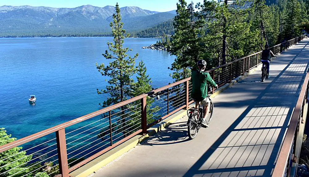 Cyclists ride along a scenic lakeside path with clear blue water and forested mountains in the background