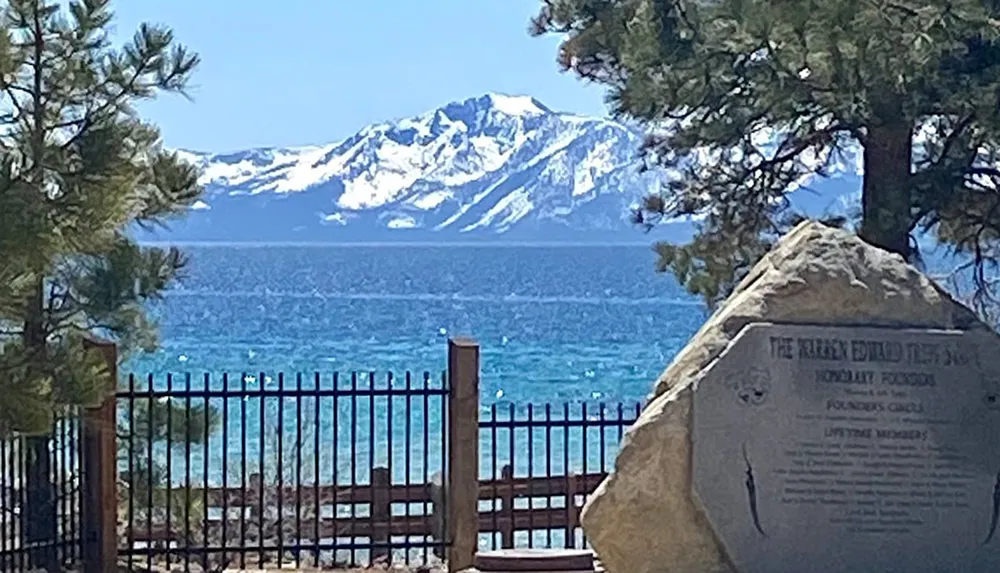 The image features a scenic view of a lake with a mountain backdrop and snow-capped peaks partially framed by trees and a fence with a commemorative rock plaque in the foreground