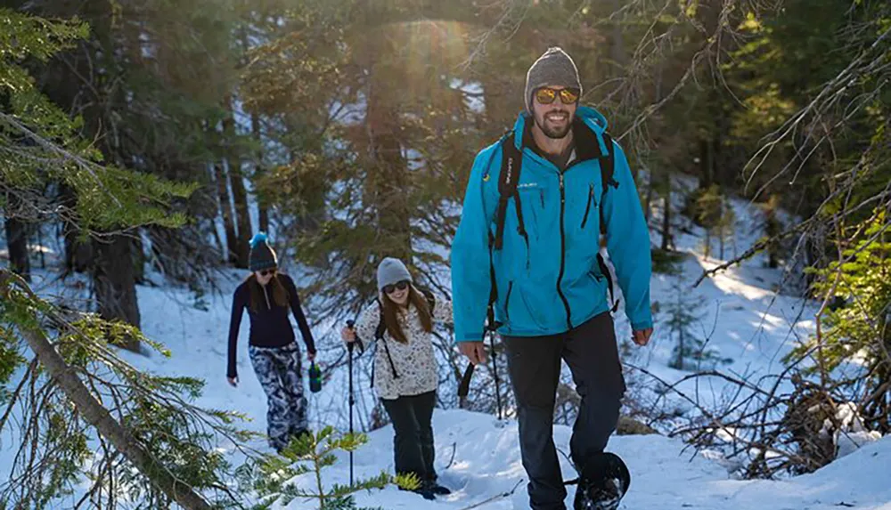 Three hikers are trekking through a snowy forest with the lead hiker smiling at the camera