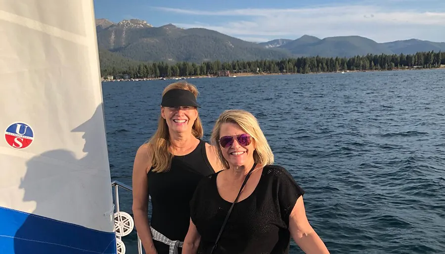 Two women are smiling on a sailboat with a picturesque backdrop of a lake and mountains.