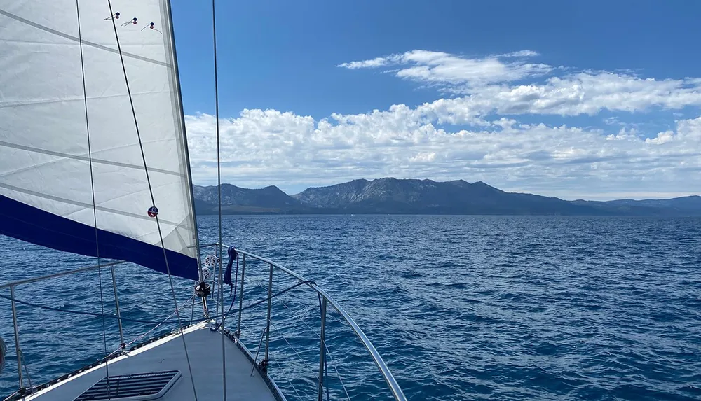 A sailboat is cruising on a calm blue sea towards a mountainous coastline under a mostly clear sky dotted with clouds