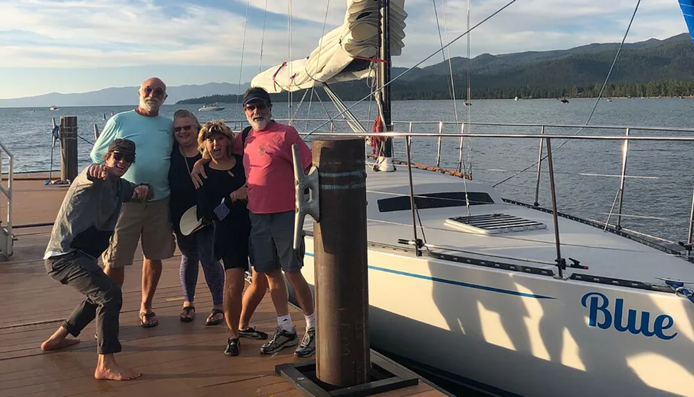 Five individuals are posing happily on a dock beside a sailboat named Blue with a scenic lake and hills in the background