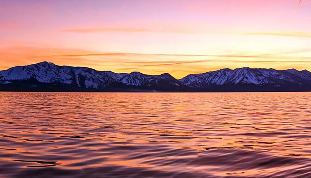 A tranquil scene of a purple and orange sunset over a calm lake with a backdrop of snow-capped mountains