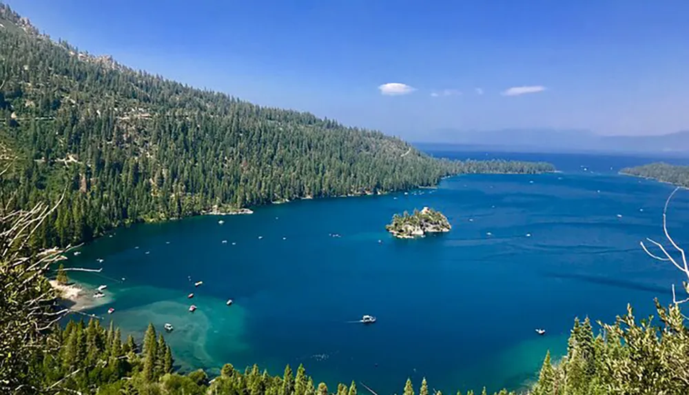 The image shows a panoramic view of a vibrant blue alpine lake surrounded by dense coniferous forests with boats on the water and a small island in the center set against a backdrop of mountains under a clear sky with few clouds