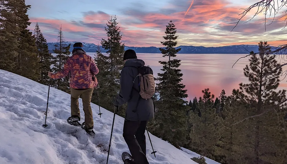 Two hikers with snowshoes are enjoying a vibrant sunset over a snowy mountain landscape and a calm lake