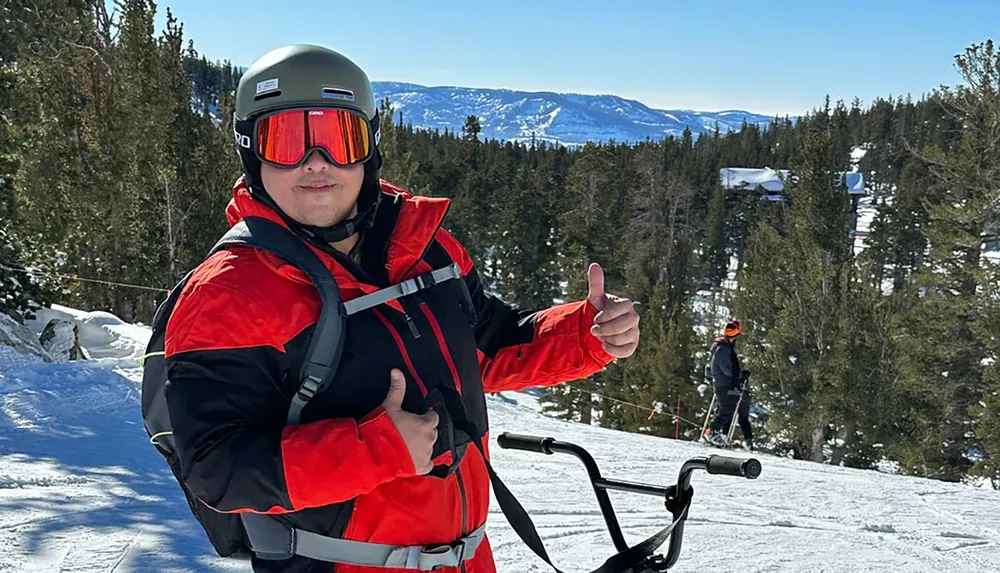 A person in ski gear gives a thumbs-up on a snowy mountain slope with skiers and a scenic mountain view in the background