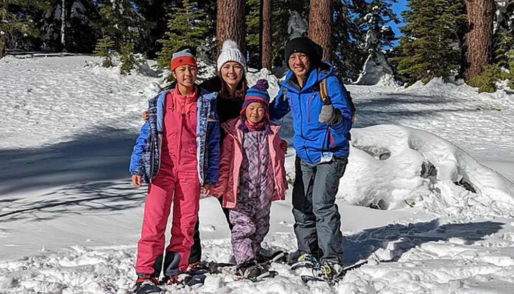 A family of four is dressed in winter attire smiling for a photo in a snowy landscape with tall pine trees in the background
