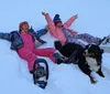 Two children and a dog are enjoying playing in the snow with one child lying down and another sitting both are wearing winter gear while the dog stands beside them