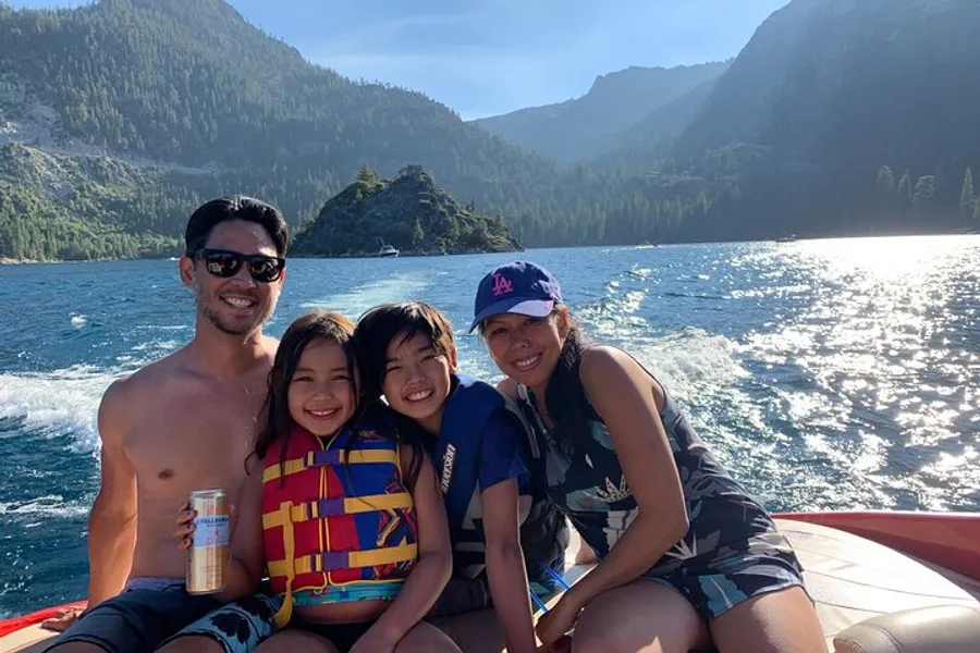 A family is smiling and enjoying a sunny boat ride on a scenic lake with mountains in the background.