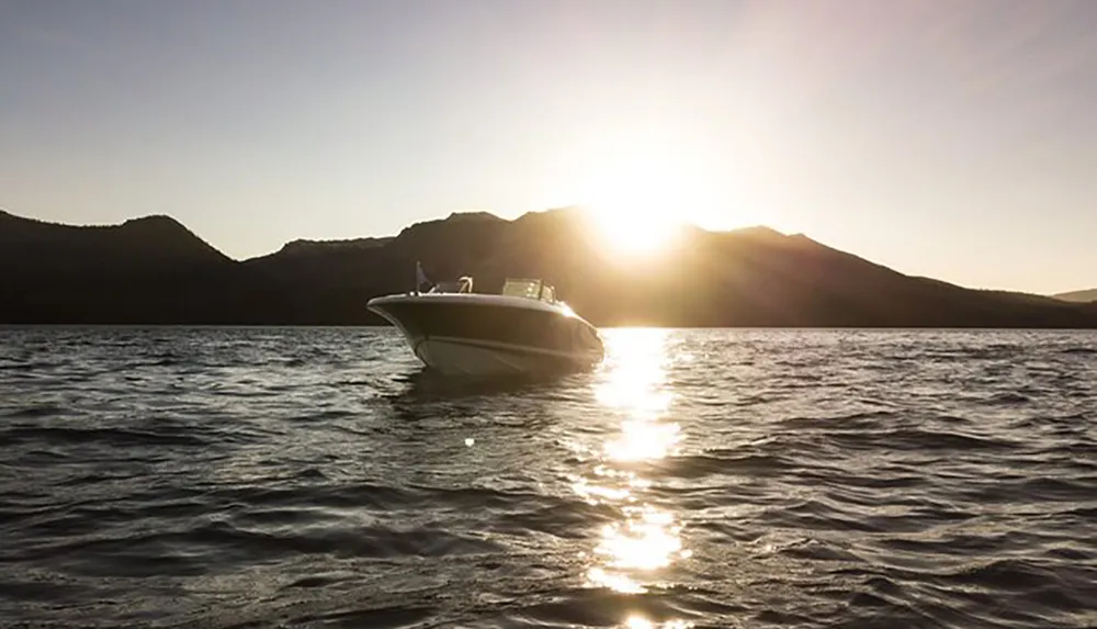 A motorboat is cruising on a tranquil lake against the backdrop of a mountain silhouette and a setting sun